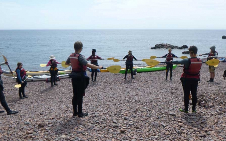 A group of people stand in a circle holding paddles in an apparent kayaking exercise. Several colorful kayaks rest on the shore with a vast lake in the background. 